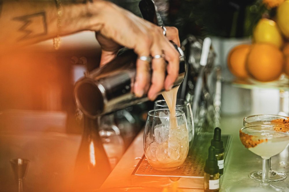 Bartender pouring a shaken cocktail into a rocks glass, with citrus and coupe glasses in the background